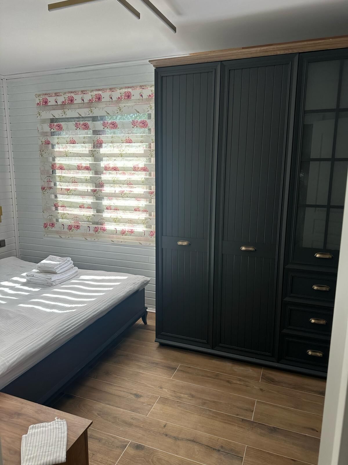A bedroom with a bed, a wardrobe, and a window with floral blinds.