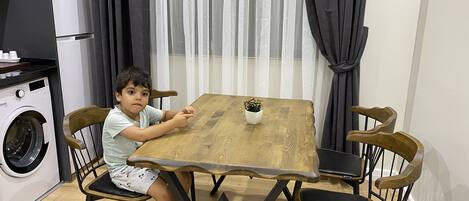 Een kind zittend aan een houten tafel met een kleine potplant, omringd door stoelen.