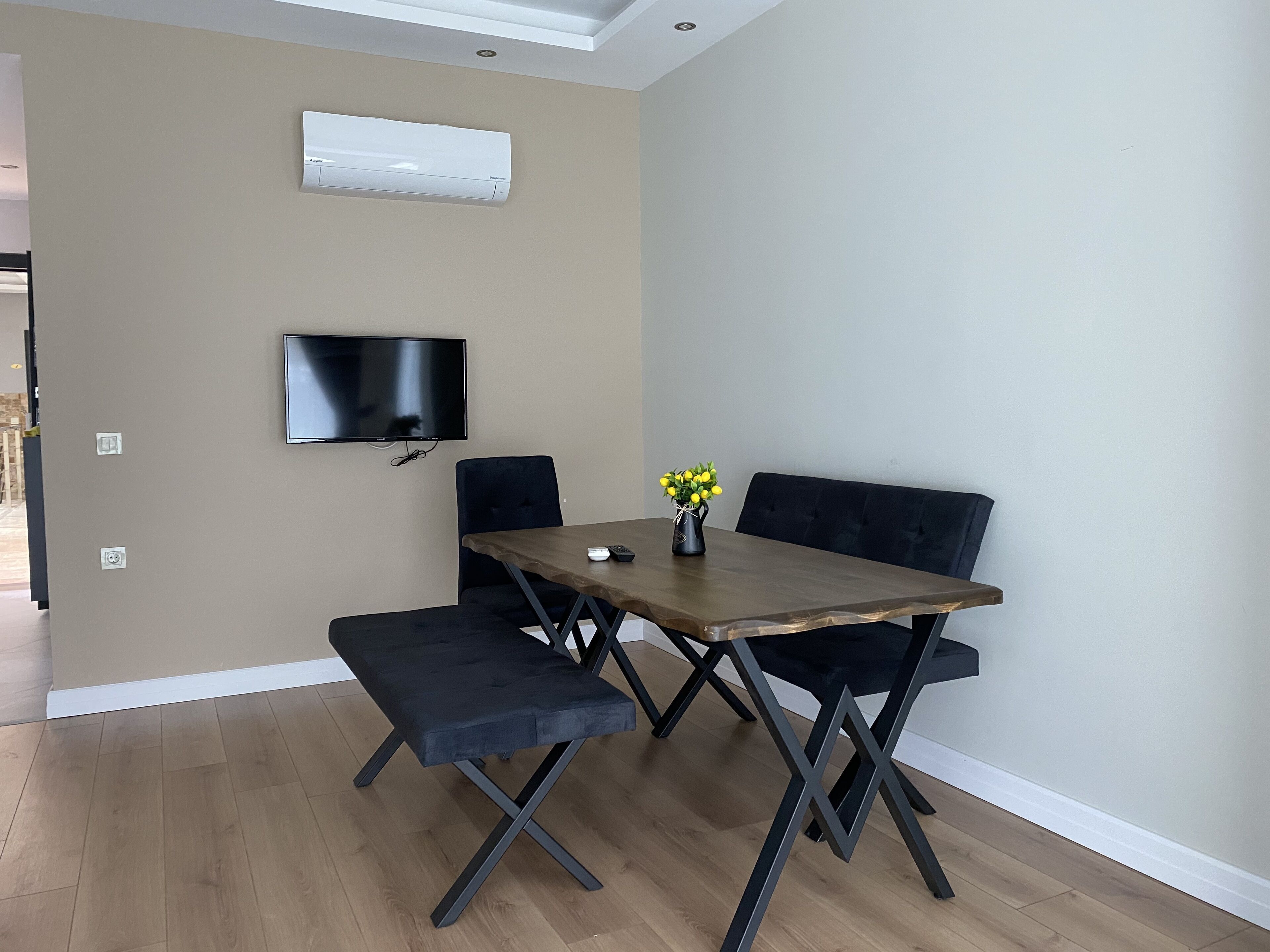 A modern dining area with a wooden table, black upholstered chairs, a bench, a flat-screen TV mounted on the wall, and a vase with yellow flowers on the table.