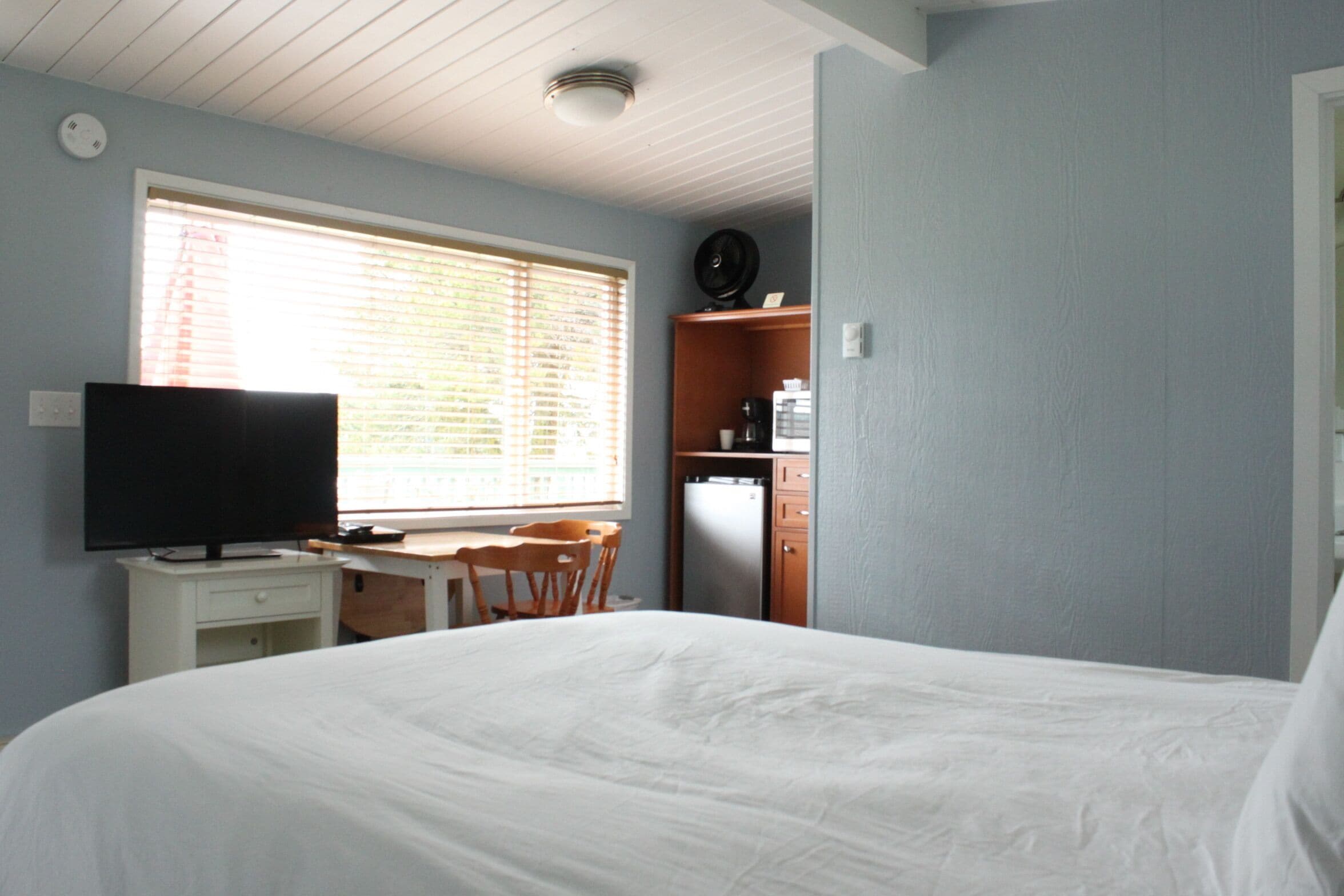 A bedroom with a bed, a desk, a chair, a television, and a small kitchenette.
