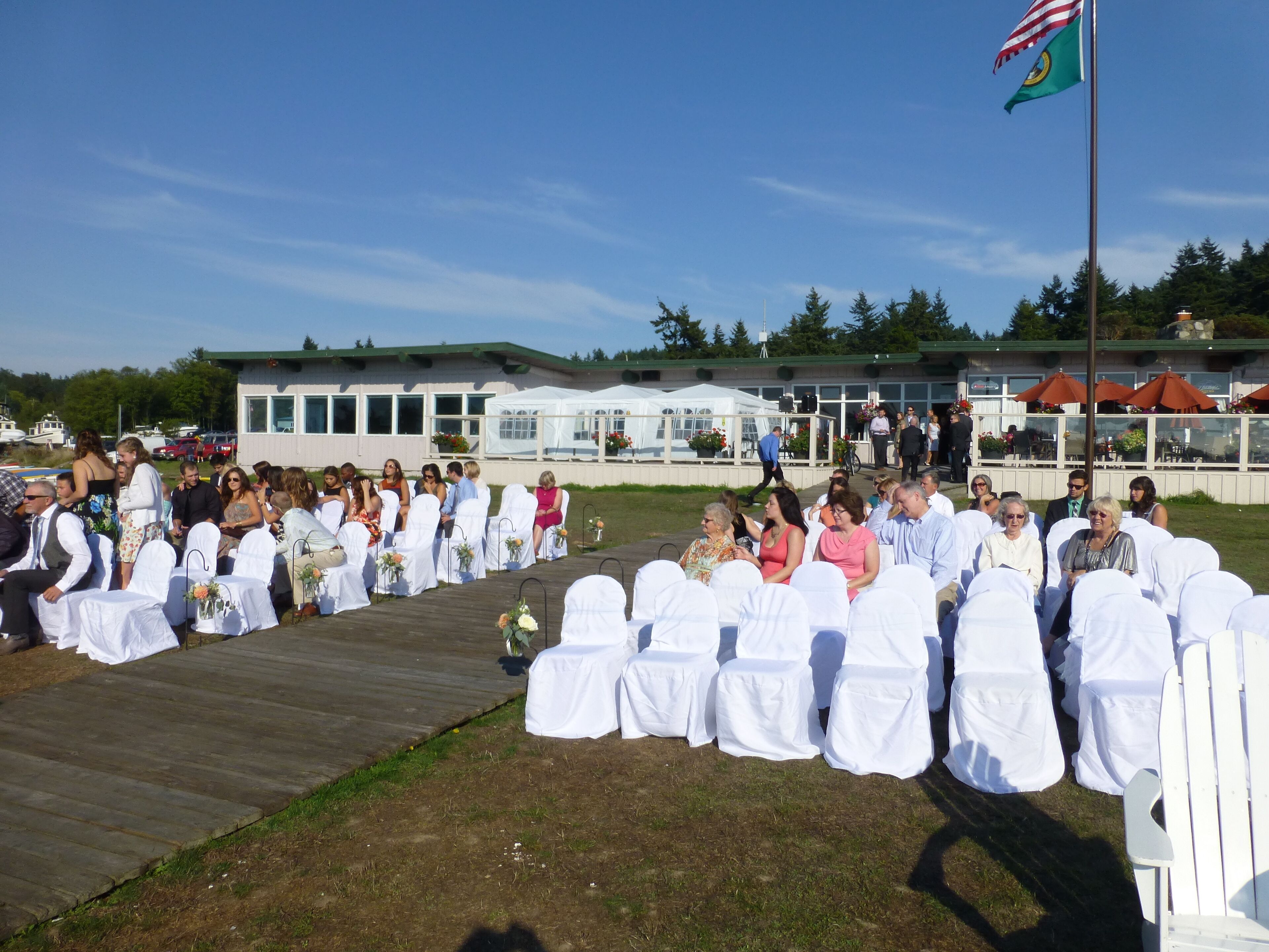 A wedding ceremony outdoors with guests seated on white chairs, a stage, and a building in the background.