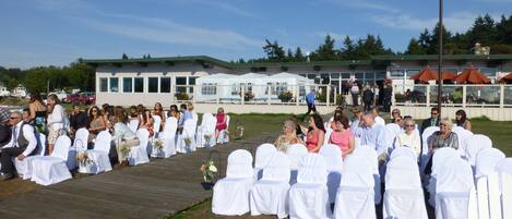 A wedding ceremony outdoors with guests seated on white chairs, a stage, and a building in the background.