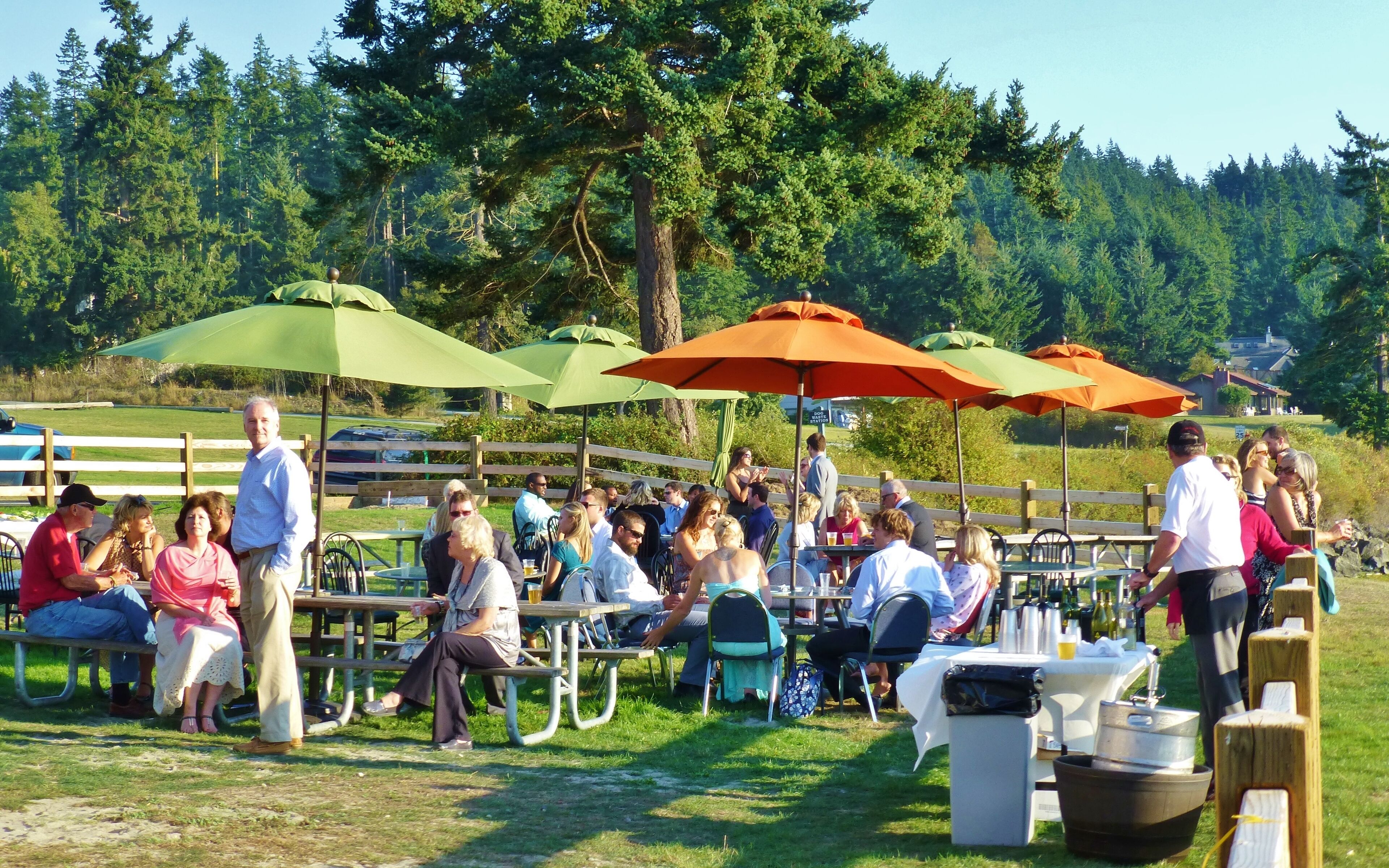 An outdoor seating area with tables and chairs under umbrellas, surrounded by a wooden fence and trees.