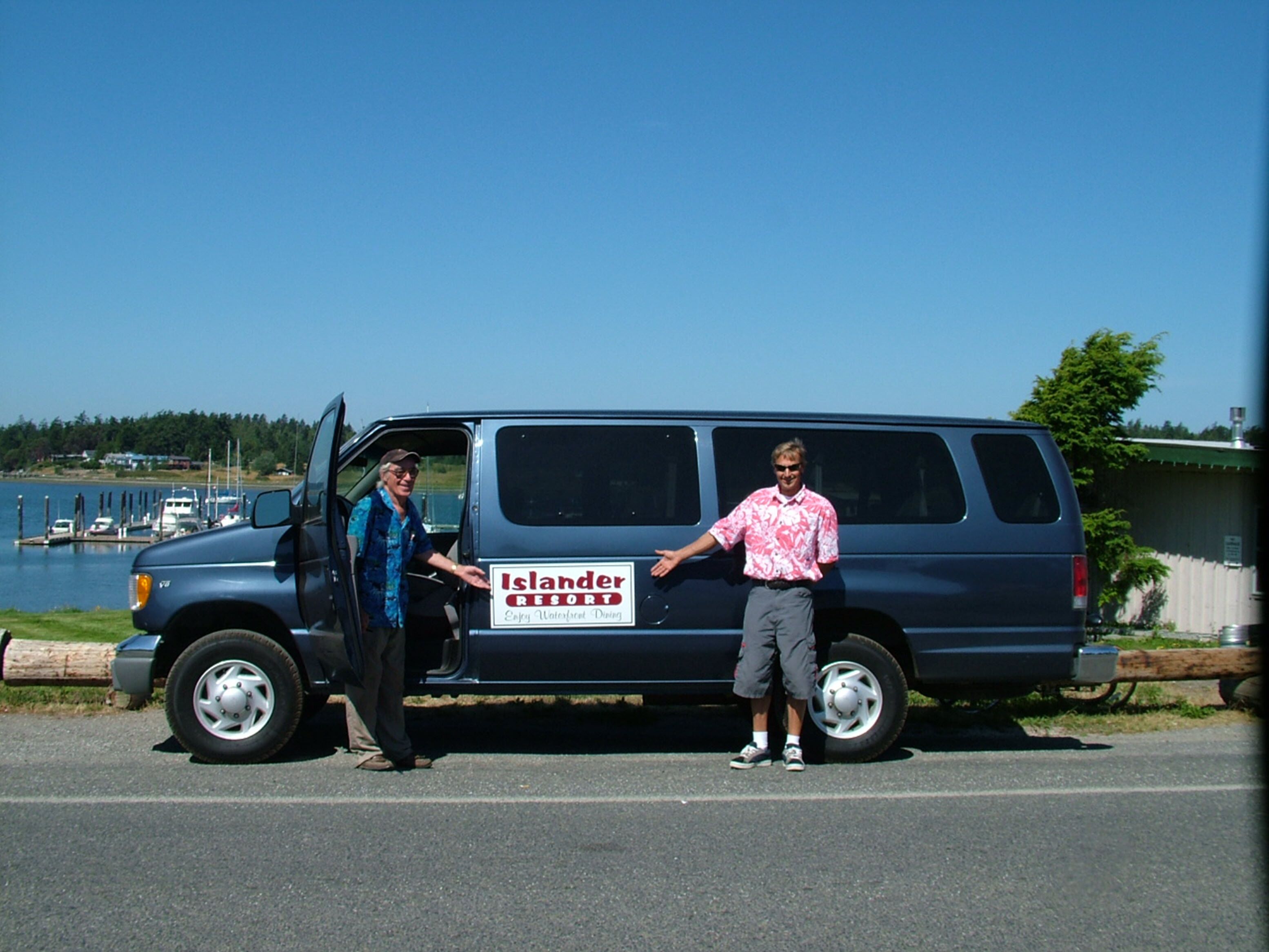 A blue van with a sign that reads "Islander Resort" parked by a road with a body of water and docked boats in the background.
