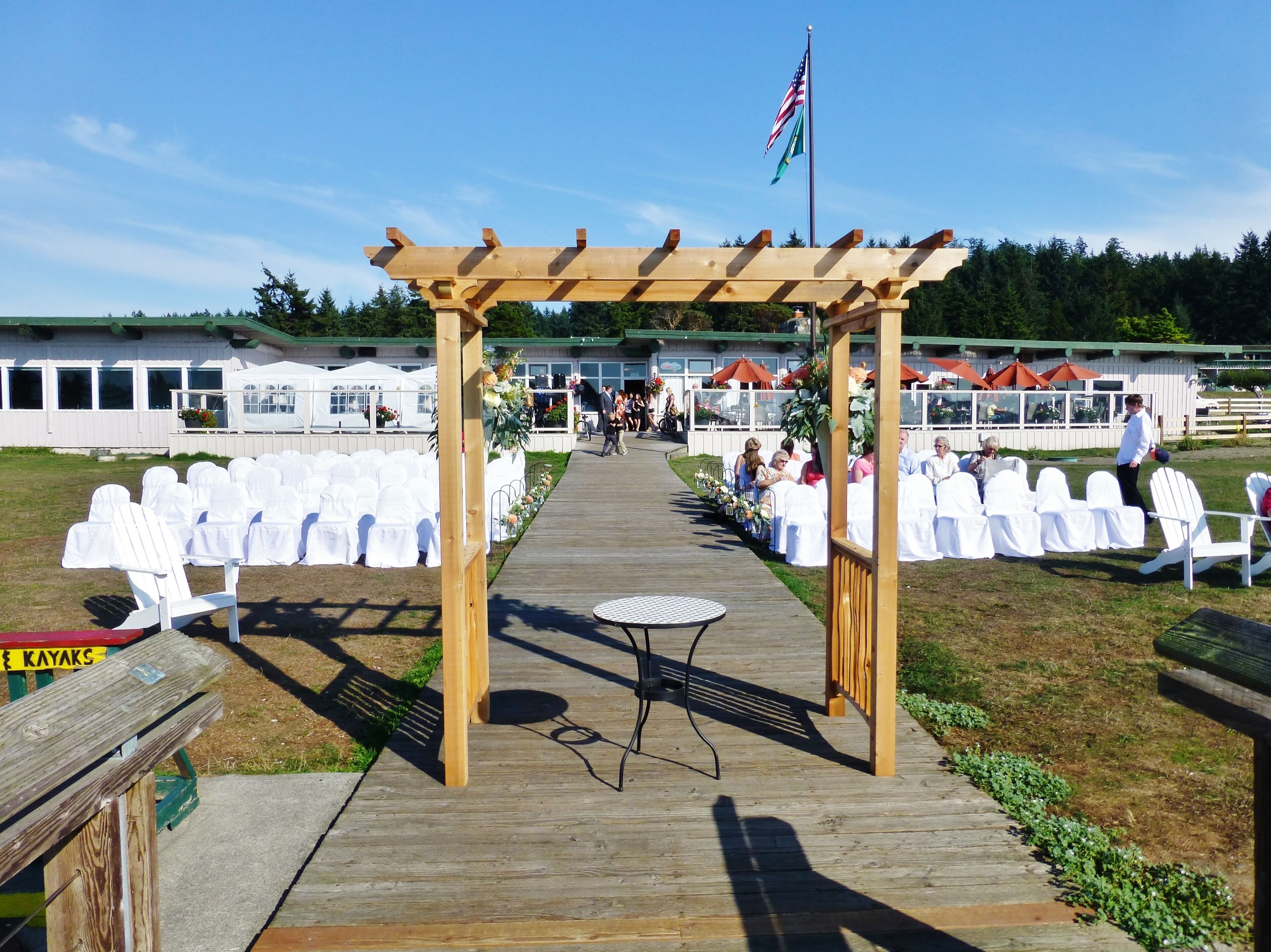 Une terrasse en bois mène à un espace abrité où se trouvent des chaises et des tables blanches, un drapeau et, au fond, un bâtiment.