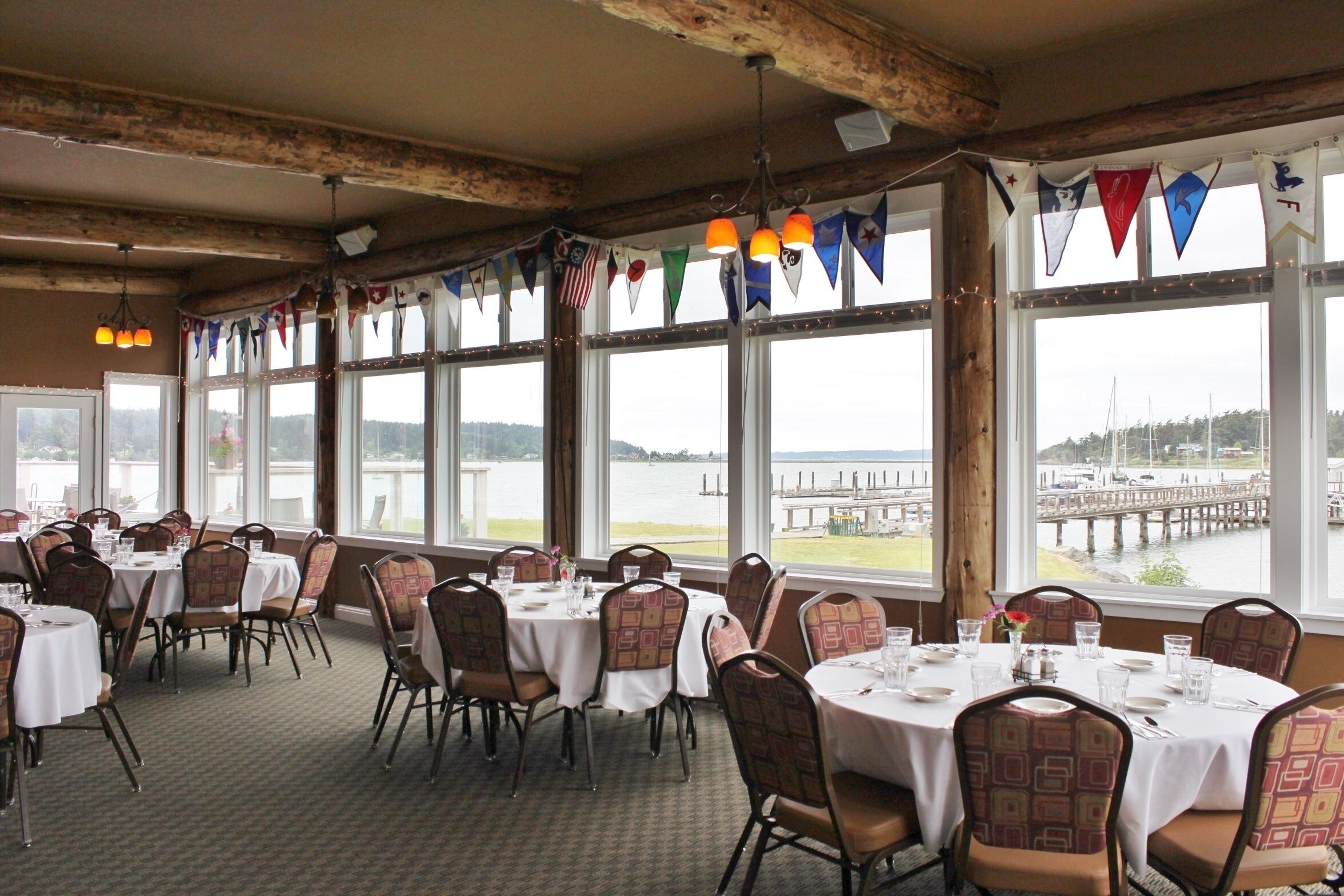 A dining area with tables set for service, large windows with a view of a body of water and dock, and colorful flags hanging from the ceiling.