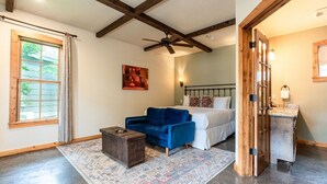 Un dormitorio con cama, sofá azul, cofre de madera, alfombra estampada, ventilador de techo y ventana con cortinas.