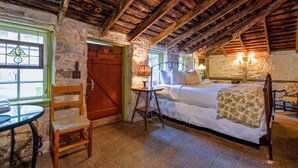 A rustic room with a wooden ceiling, a bed with a floral bedspread, a stone wall, and a small table with a chair.
