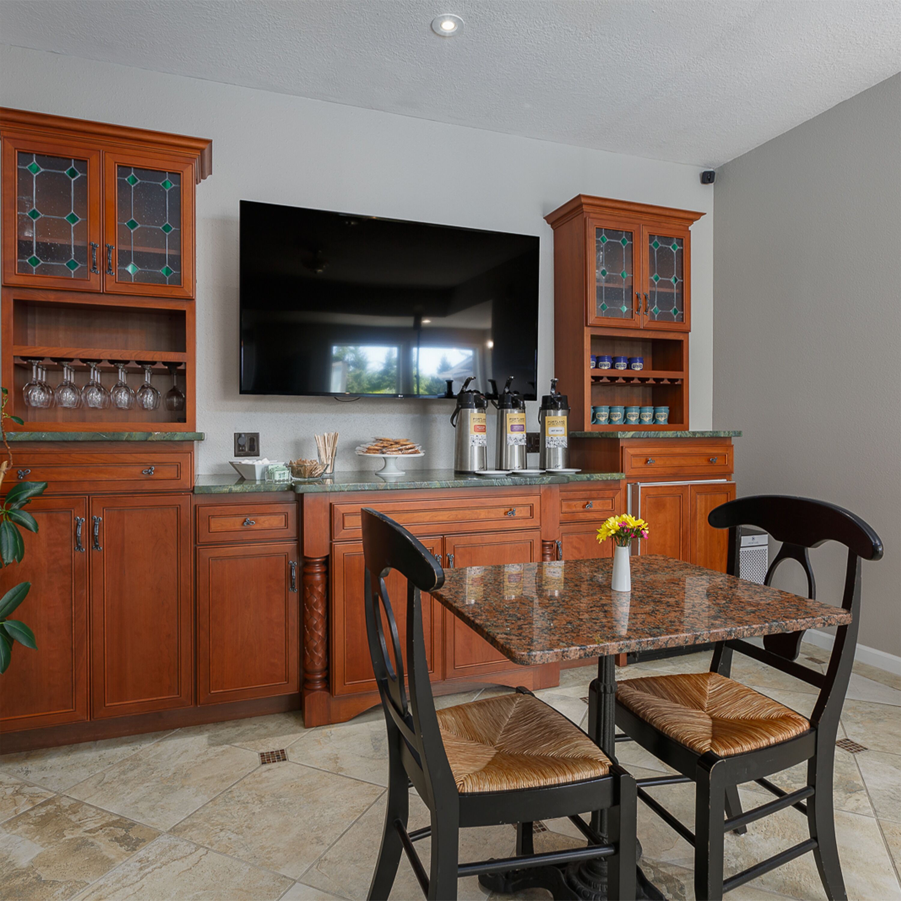 A kitchen with wooden cabinets, a granite countertop, a large flat-screen TV, and two chairs with woven seats.