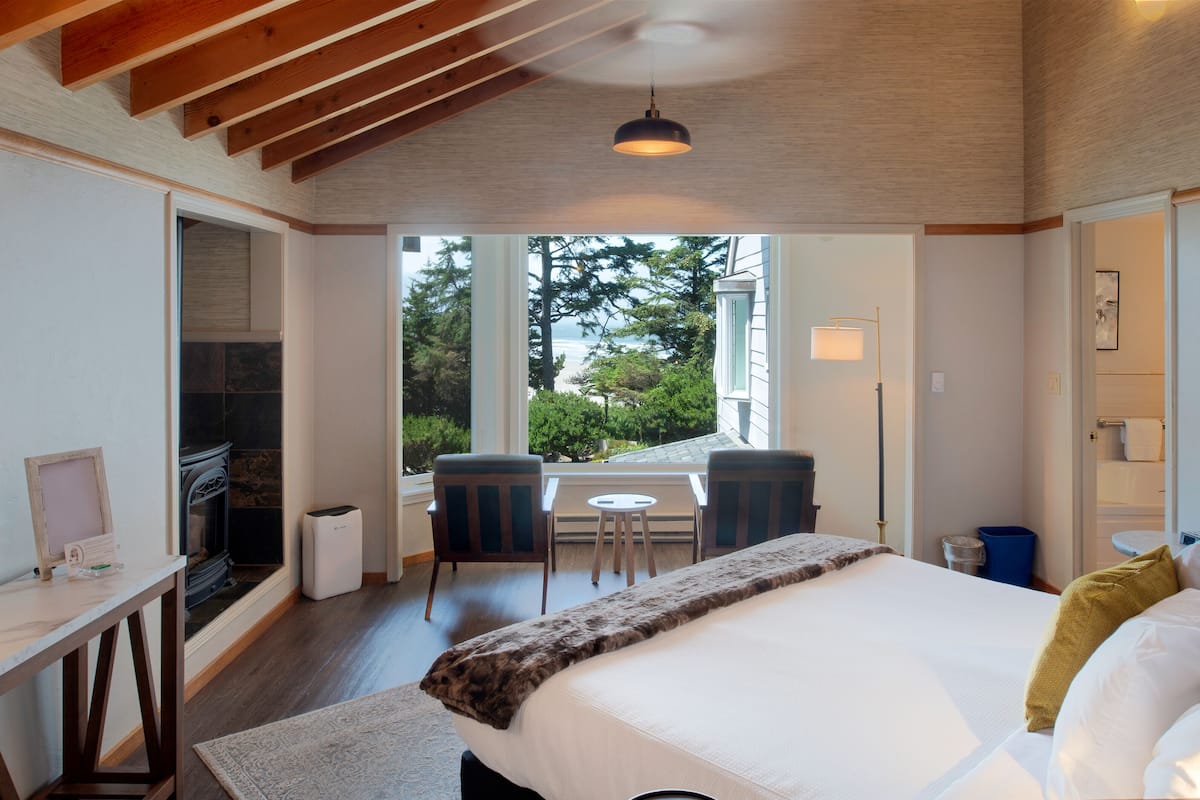 A bedroom with a large bed, a fireplace, a dining area with chairs, and a view of the outdoors.