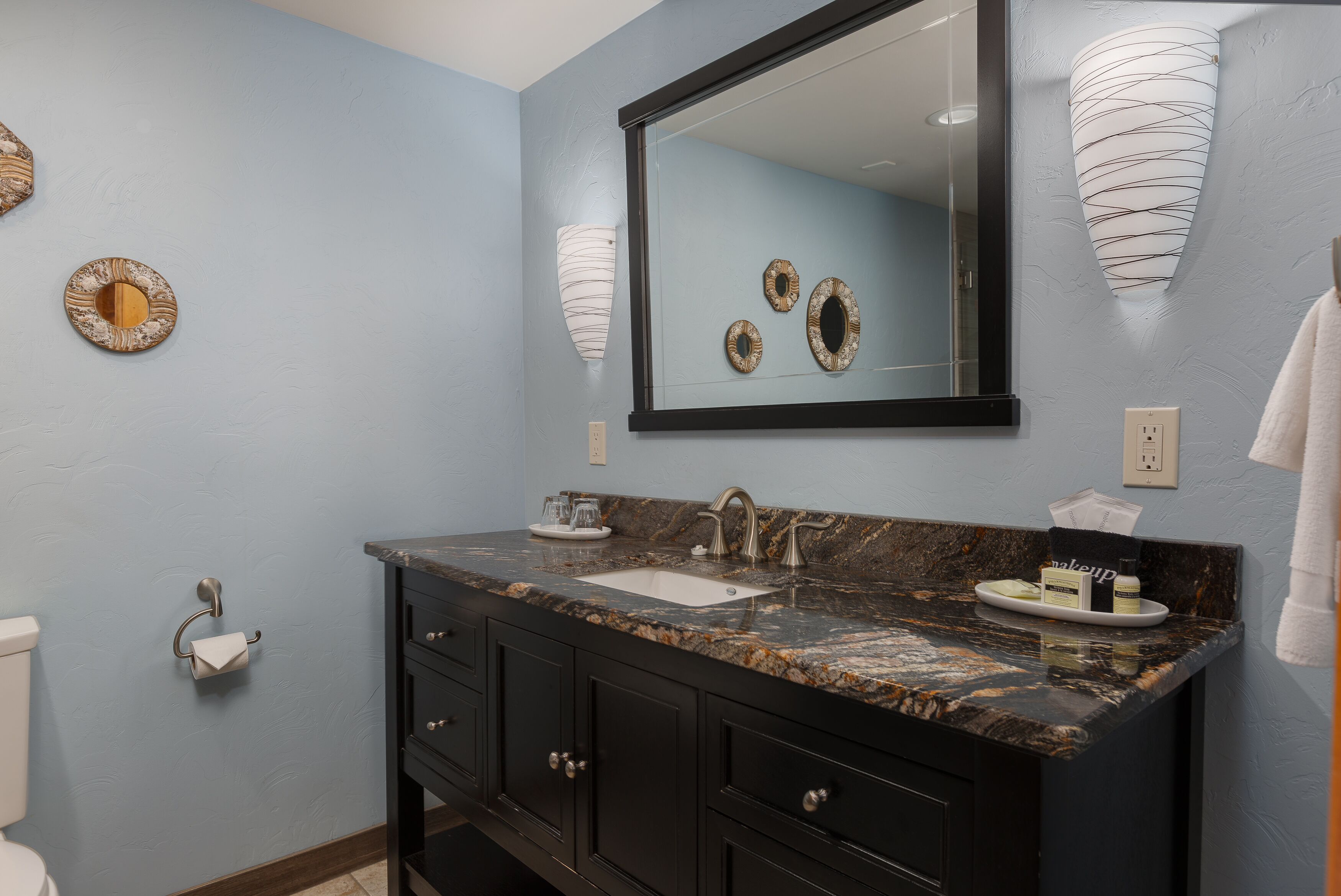 A bathroom with a marble sink, dark cabinets, a mirror, and decorative wall sconces.