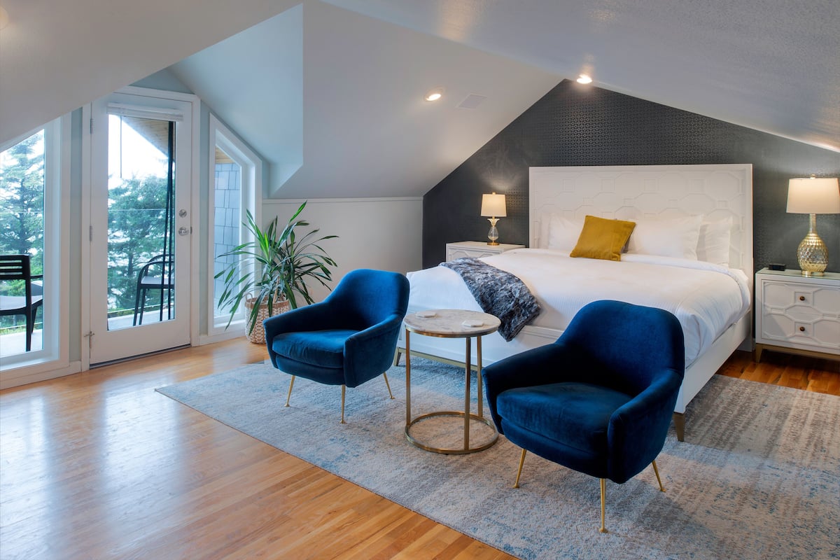 A modern bedroom with a large bed, two blue armchairs, a small round table, and a large window with a view of trees.