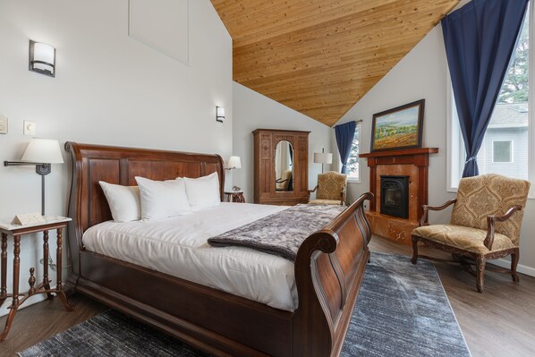 A bedroom with a wooden bed, a nightstand, a lamp, a chair, a mirror, and a fireplace.