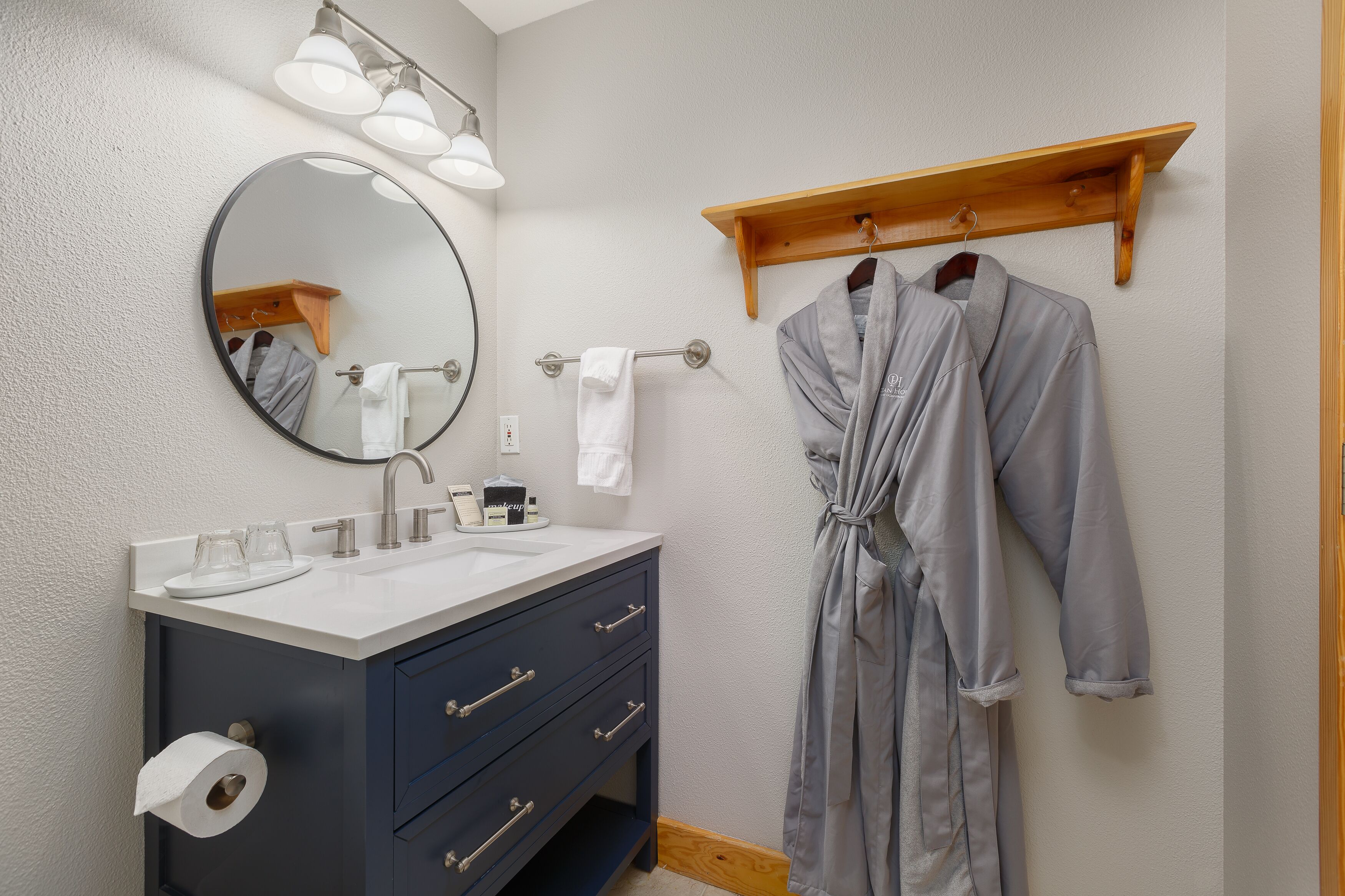 A bathroom with a round mirror, a sink, a towel rack, and a robe hanging on a wooden shelf.