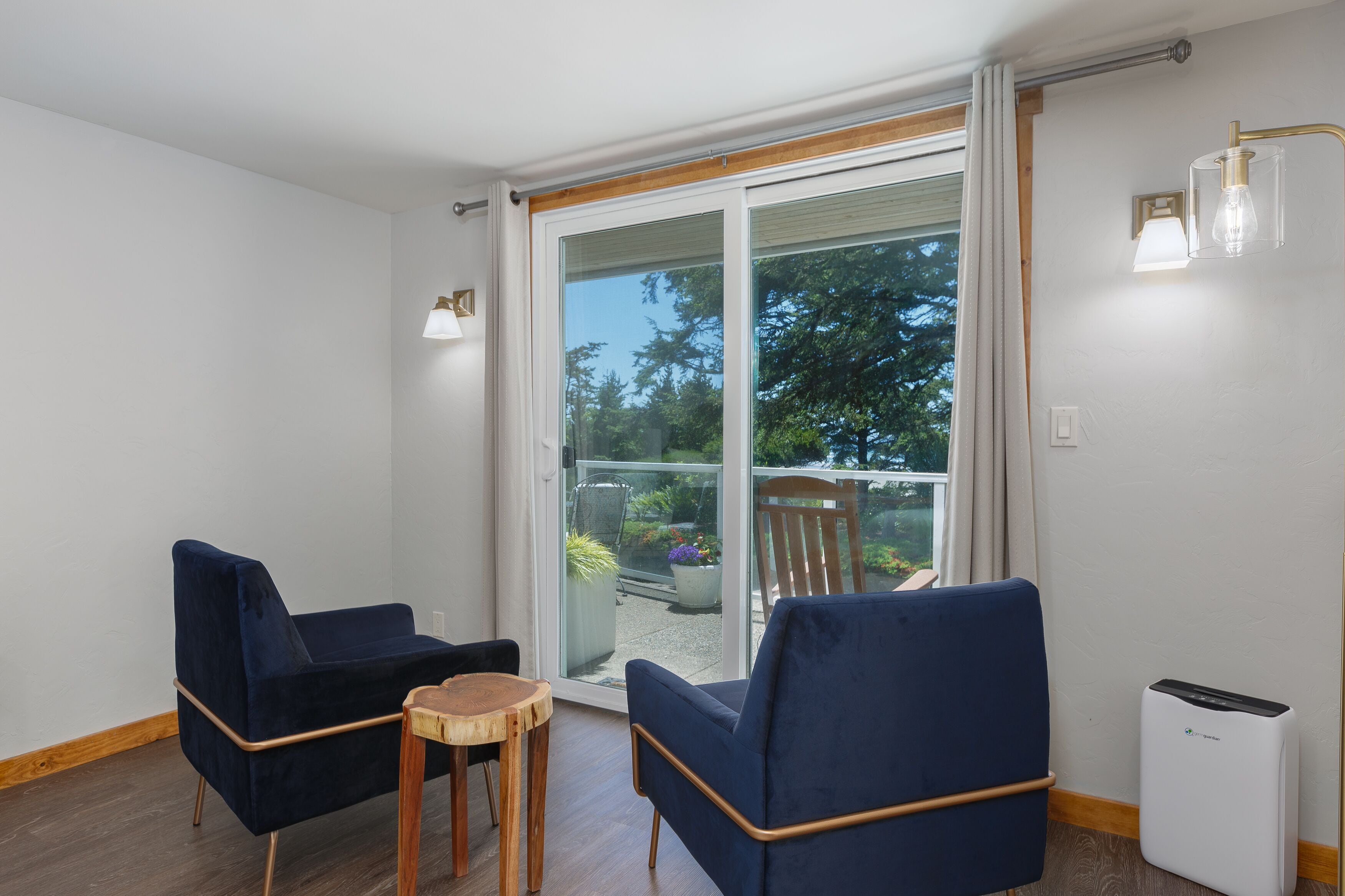Ein modernes Zimmer mit zwei blauen Sesseln, einem kleinen Holztisch und einer Schiebeglastür, die zu einem Außenbereich mit Pflanzen und einem Stuhl führt.