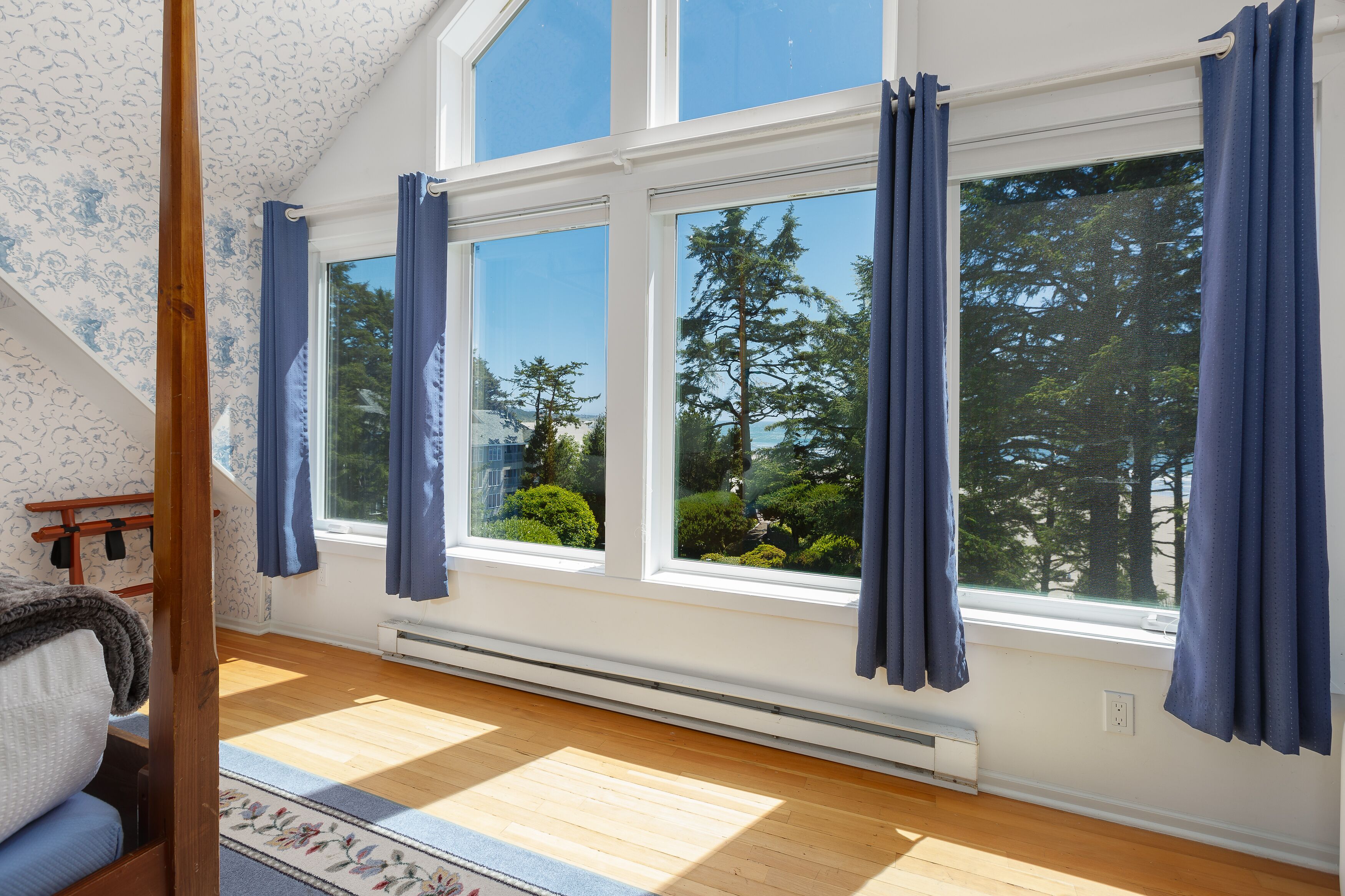 A room with a sloped ceiling, large windows with blue curtains, a wooden floor, and a view of trees and greenery outside.