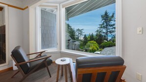 A corner room with a large window, two chairs, a small table, and a view of trees and a house. - Ocean House (Newport)