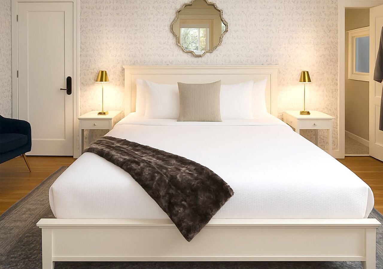 A neatly made bed with white linens and a dark throw blanket, flanked by two bedside tables with lamps, in a room with patterned wallpaper and a mirror above the bed.