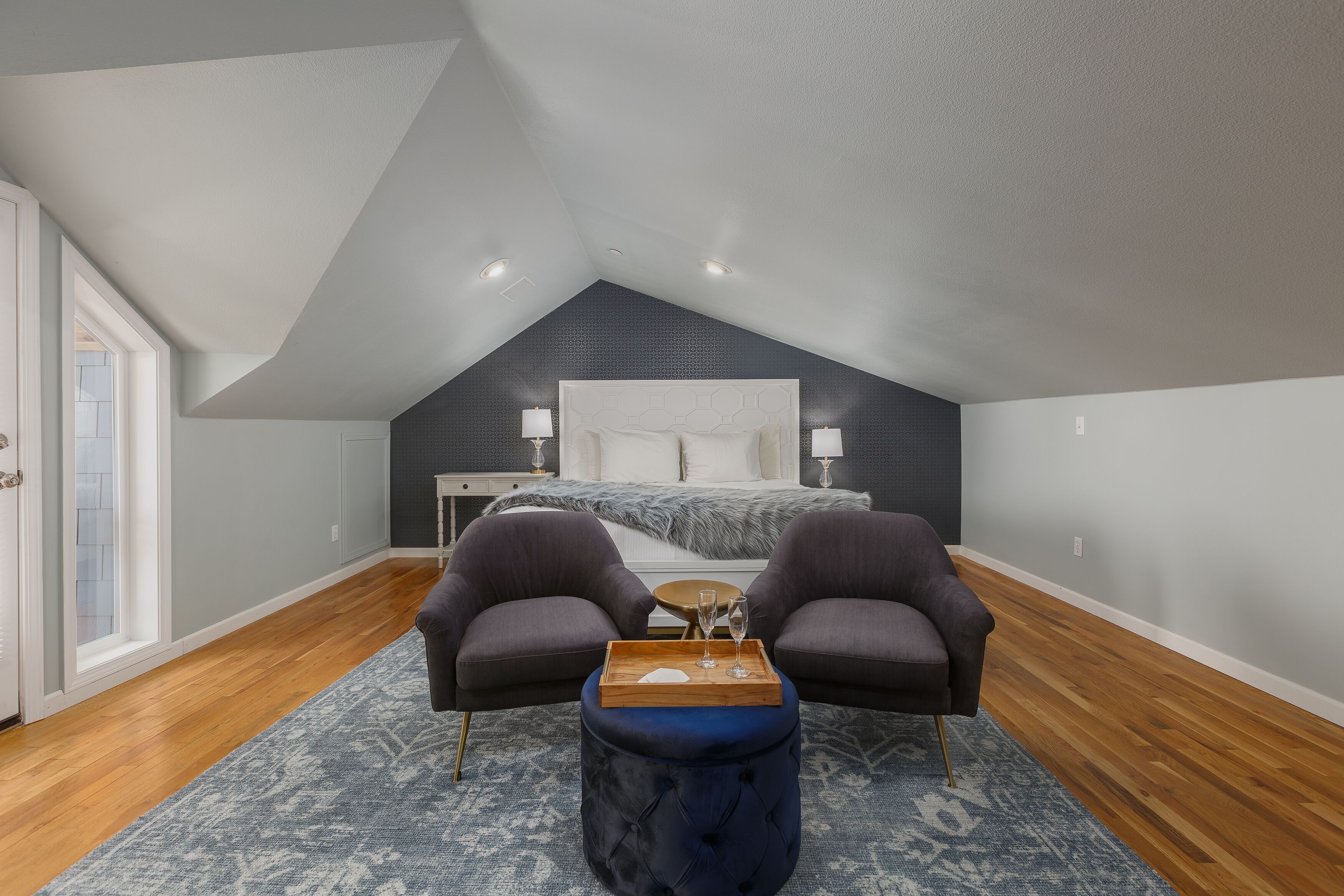Un dormitorio con techo inclinado, una cama con cabecera blanca, dos sillones oscuros, una pequeña mesa de madera con bandeja y un otoman azul.