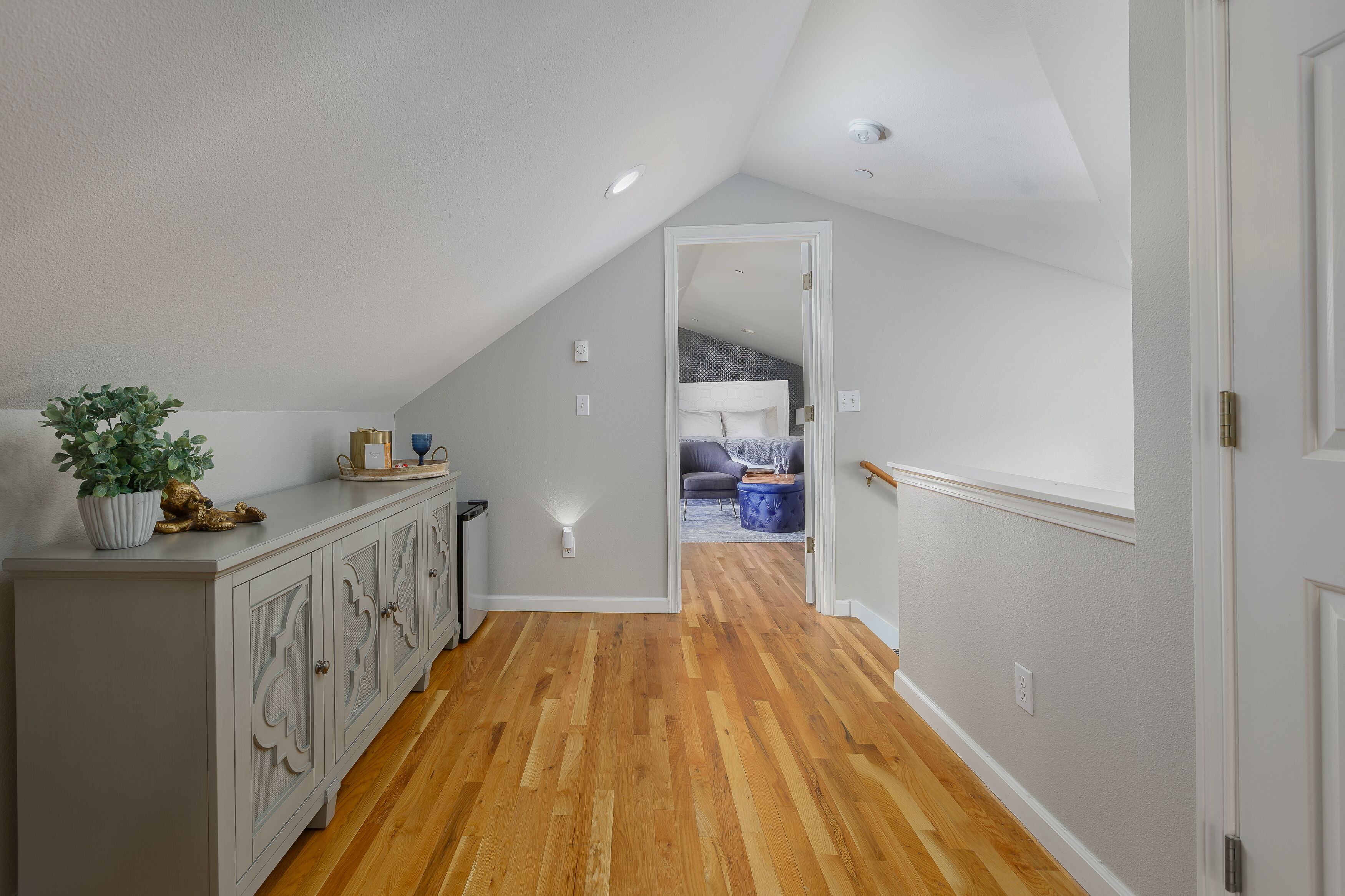 A hallway with wooden flooring, a white wall, and a door leading to a bedroom with a bed and a blue chair.