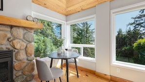 A room with a stone fireplace, a round table with chairs, and large windows offering a view of trees. - Ocean House (Newport)