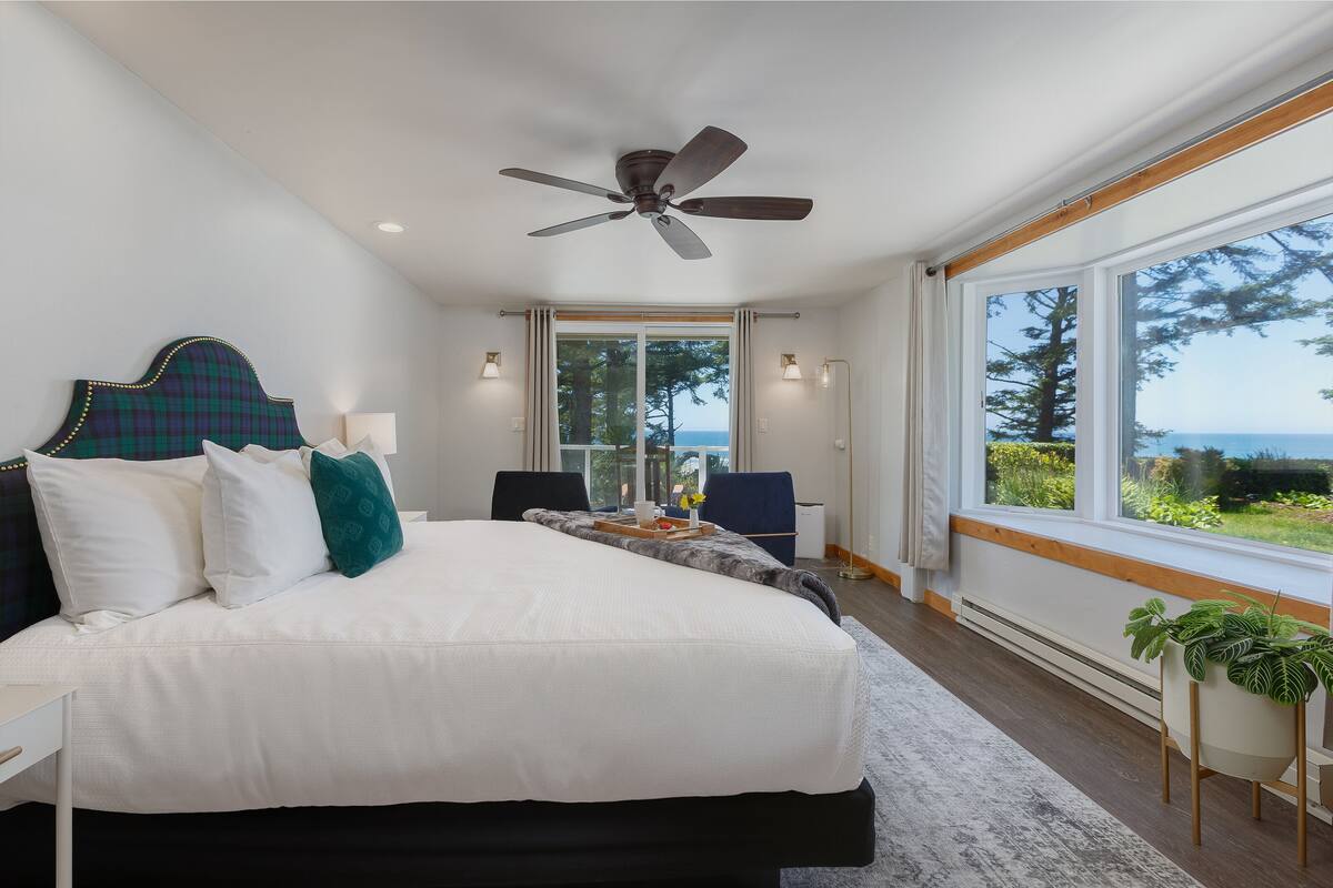 A bedroom with a bed, a ceiling fan, a large window with a view of the outdoors, and a bedside table.