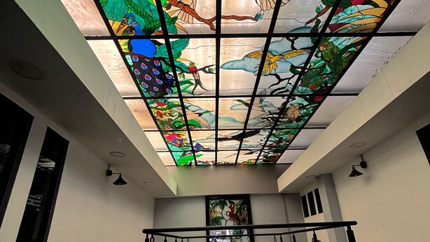 A room with a high ceiling, a skylight with stained glass, a green sofa, a potted plant, and a metal railing. - TJ Border Suites by Hosted by Me (Tijuana)