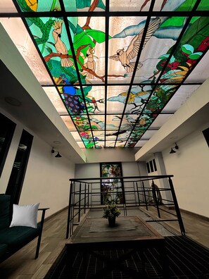 A room with a high ceiling, a skylight with stained glass, a green sofa, a potted plant, and a metal railing. - TJ Border Suites by Hosted by Me (Tijuana)
