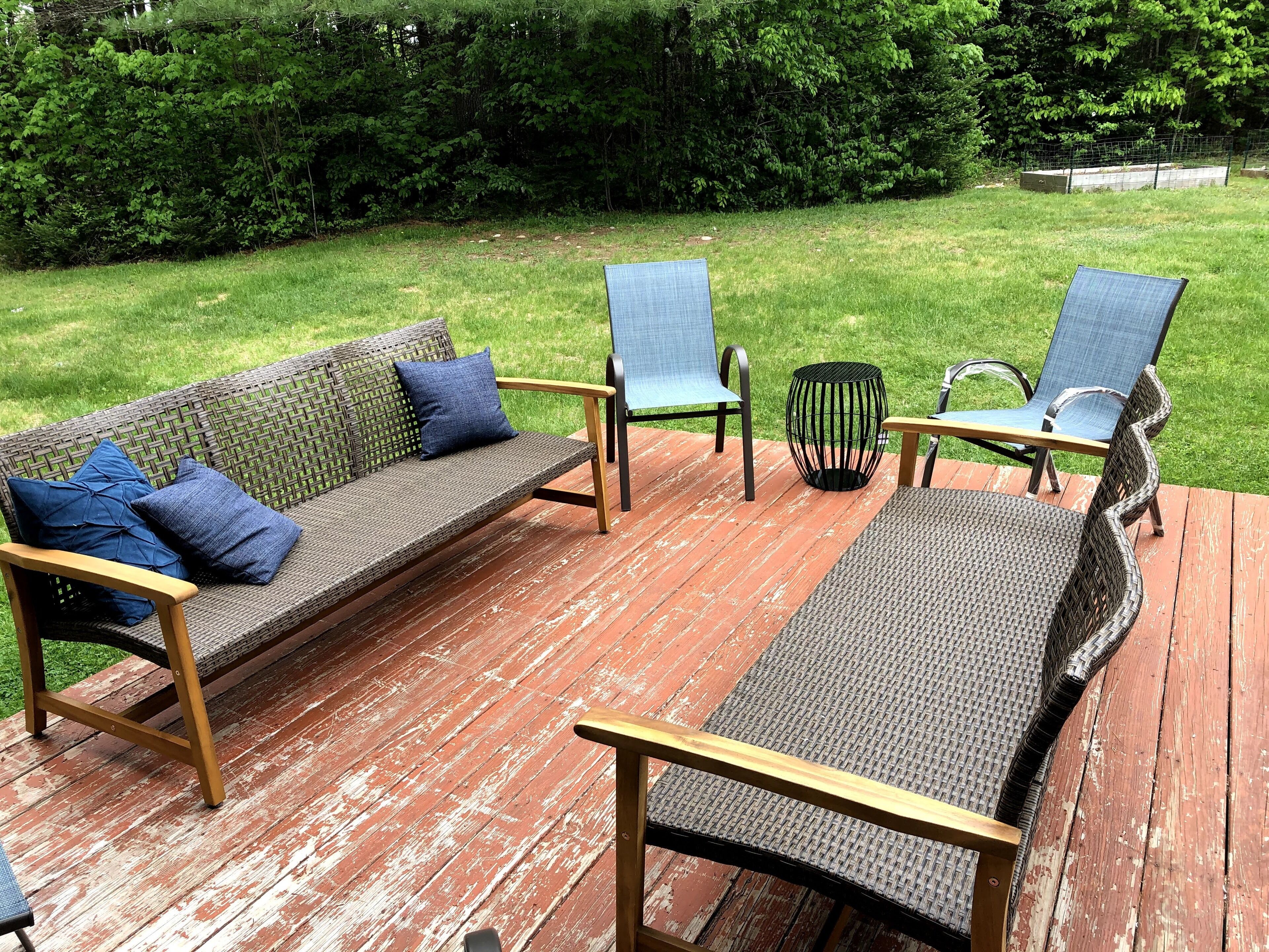 Een houten terras met een rieten bank en twee stoelen, omgeven door een gazon en dichte bomen.