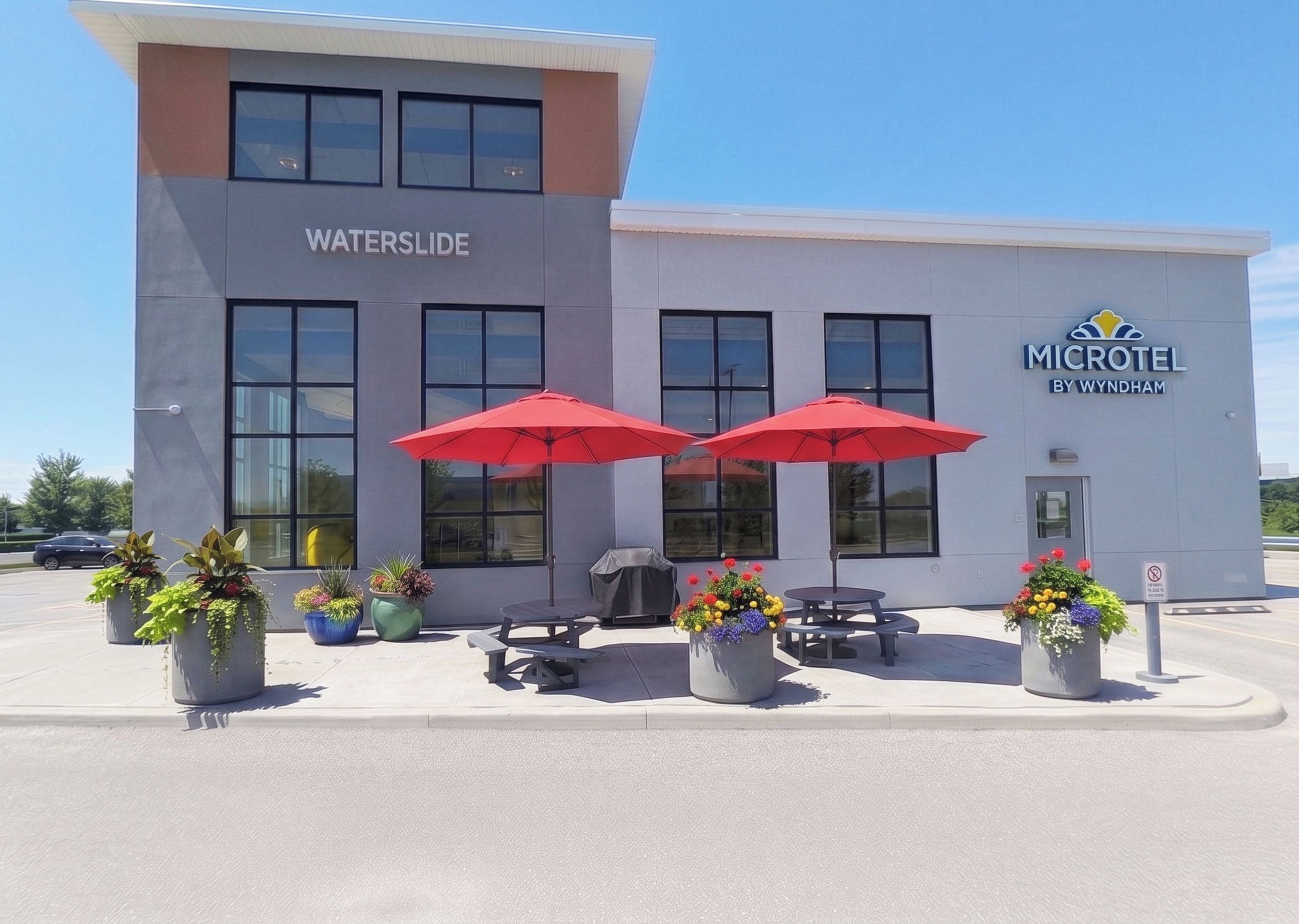 A building with the sign 'WATERSLIDE' and 'MICROTEL BY WYNDHAM' on the facade. There are outdoor tables and chairs with red umbrellas, and potted plants around.