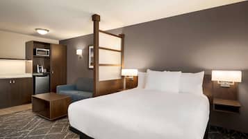 Habitación de hotel con una cama grande, un escritorio, una silla, microondas, refrigerador y una pequeña cocina.