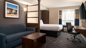 Habitación de hotel con cama, sofá azul, mesa de centro de madera, escritorio con silla, televisor y ventana con cortinas.