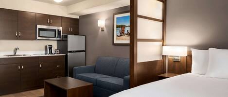 Habitación de hotel con cama, sofá, mesa de centro, microondas, refrigerador, lavamanos y una imagen de un lago.