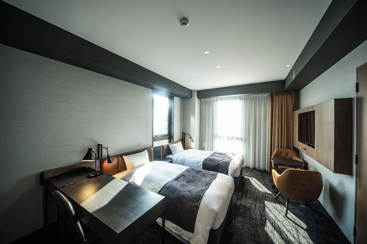 A hotel room with two beds, a desk, a chair, a TV, and a window with curtains.