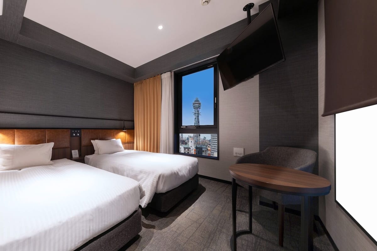 A hotel room with two beds, a TV, a window with a city view, and a small table.