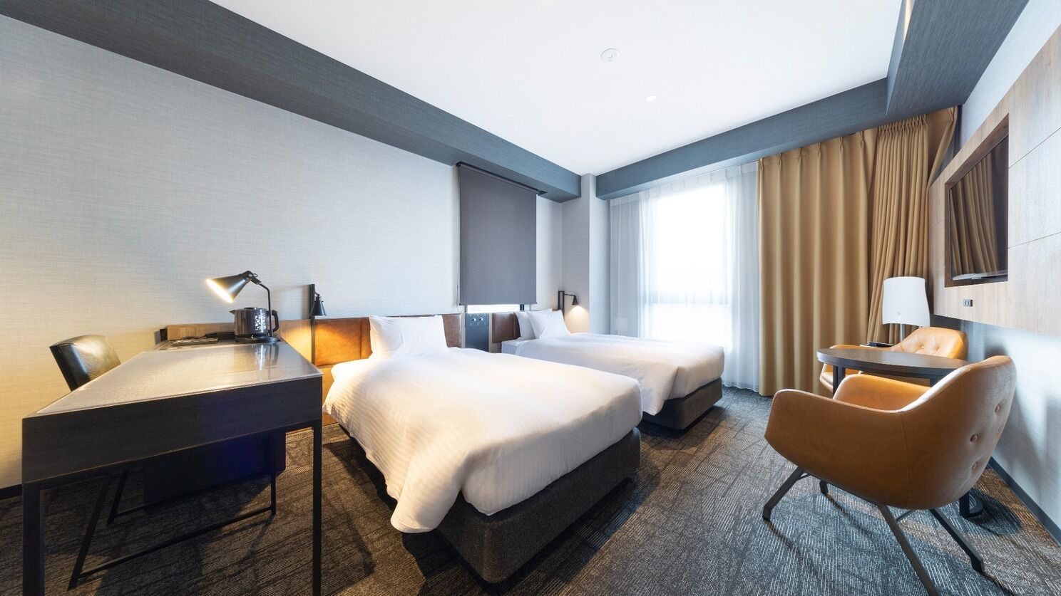 A hotel room with a desk, two beds, a chair, and a large window with curtains.