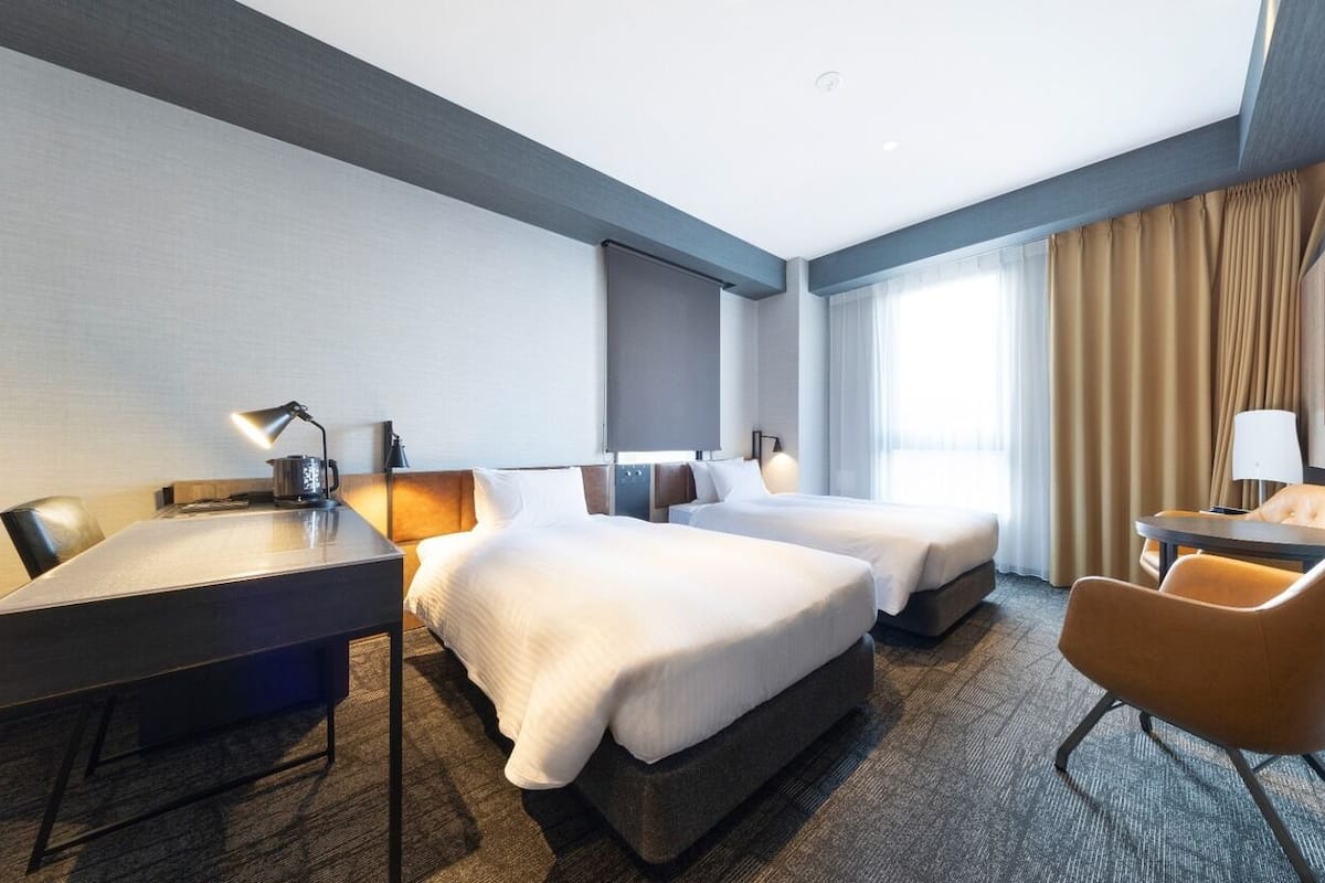 A hotel room with a desk, two beds, a chair, and a large window with curtains.