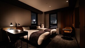 Habitación de hotel con dos camas, un escritorio, una silla y vista a la ciudad de noche.