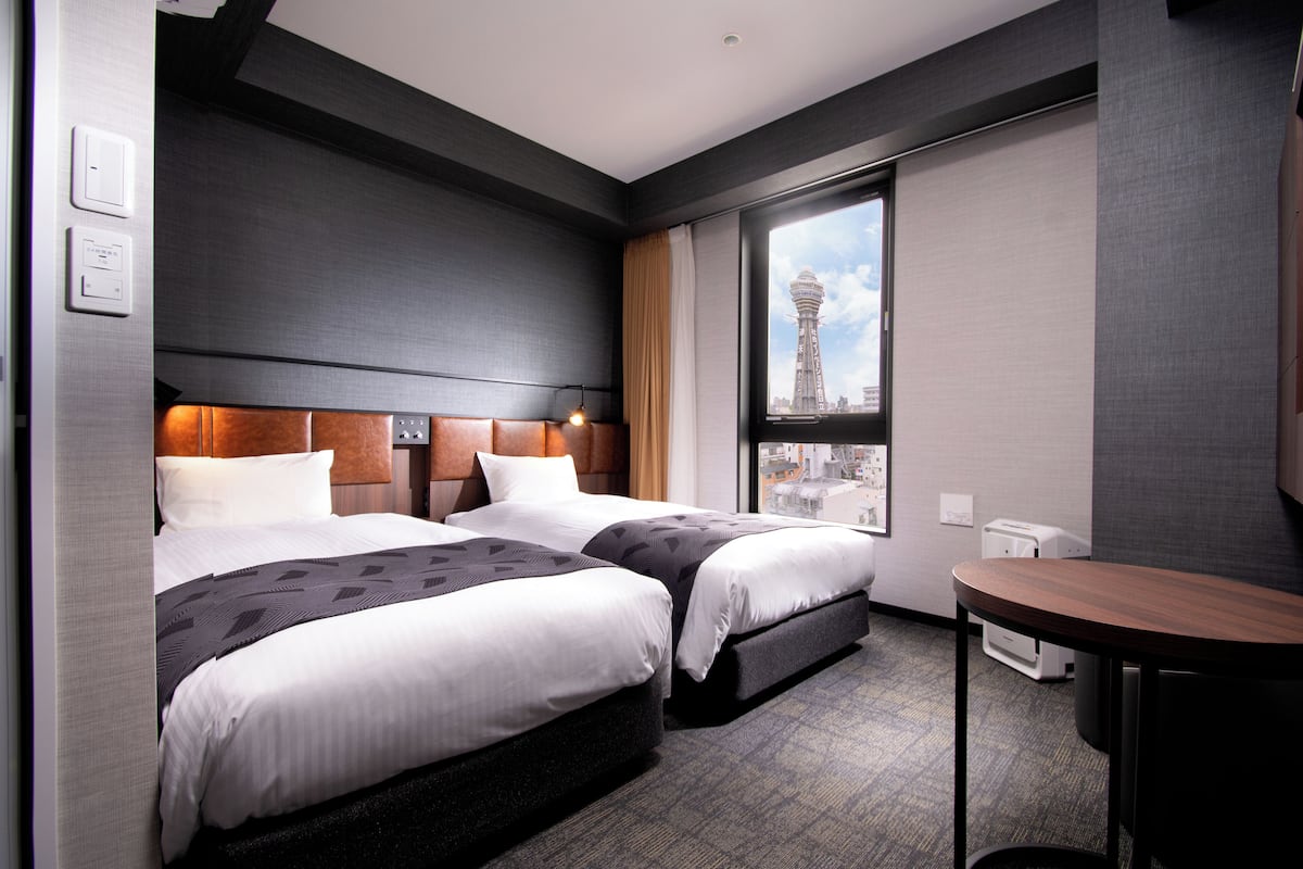 A hotel room with two beds, a small table, and a view of a tower through a window.