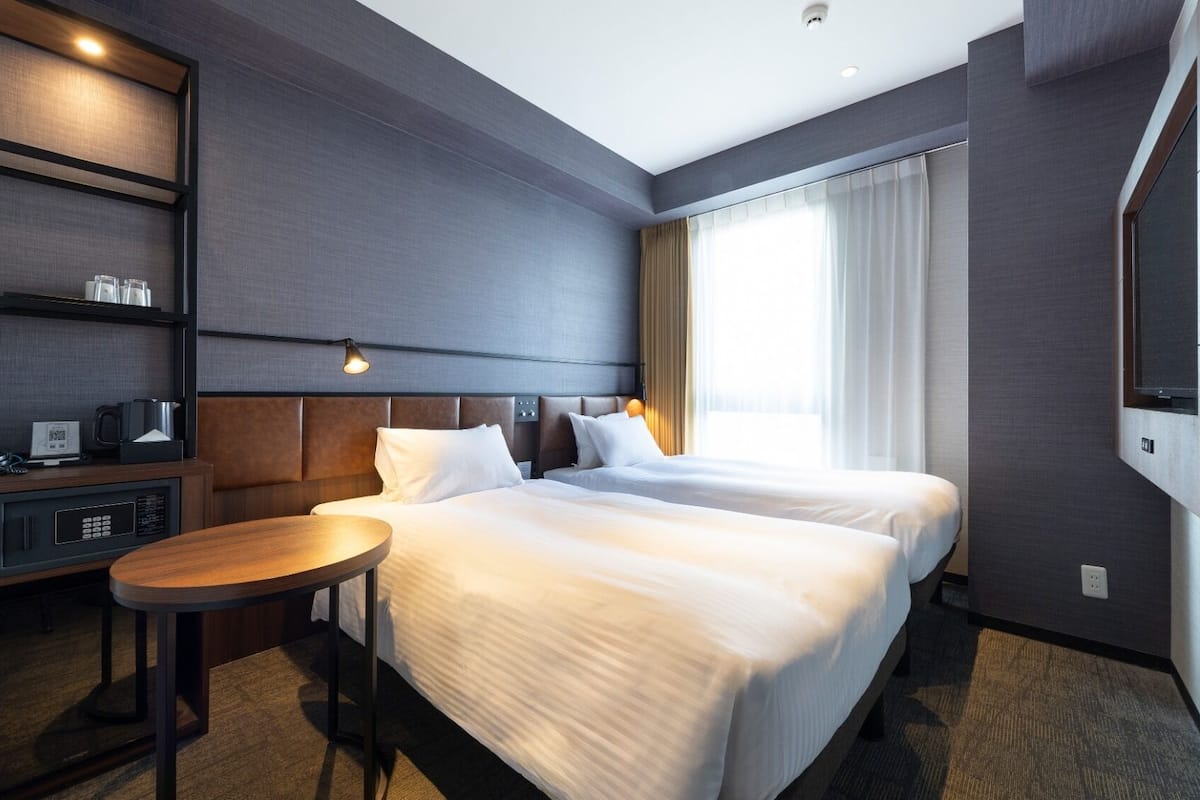 A hotel room with two beds, a desk with a chair, a TV, and a minibar.