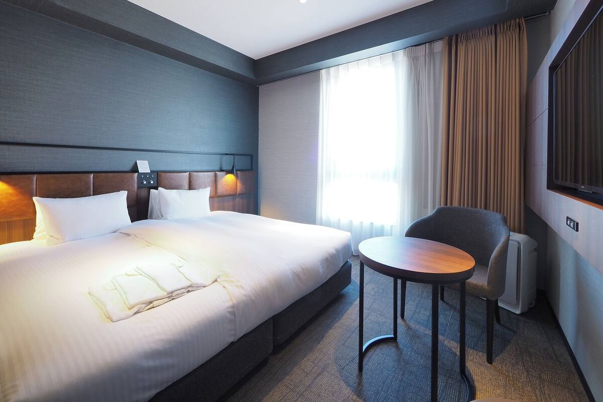 A hotel room with a large bed, a small round table, a chair, a bedside lamp, and a large window with curtains.