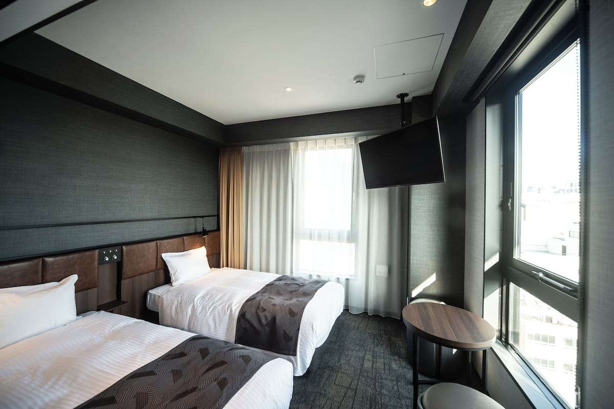 A hotel room with two beds, a TV, a small table, and a window with curtains.