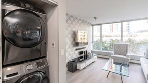 Een modern appartement met een ingebouwde wasmachine en droger, een glazen salontafel, een witte bank en een groot raam met uitzicht op de stad.