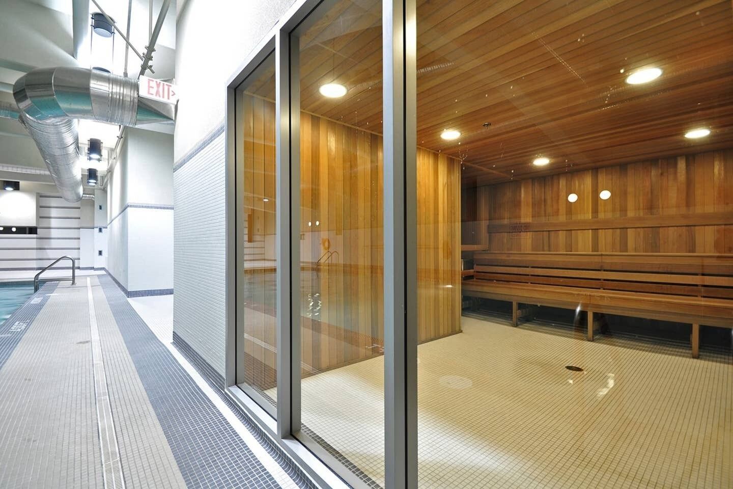 A sauna with wooden benches and a glass wall, a pool area with a tiled floor, and an exit sign.