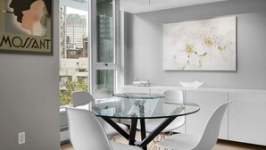 Een moderne eetruimte met een glazen tafel en witte stoelen, een groot bloemschilderij aan de muur, en een uitzicht op gebouwen door een groot raam.