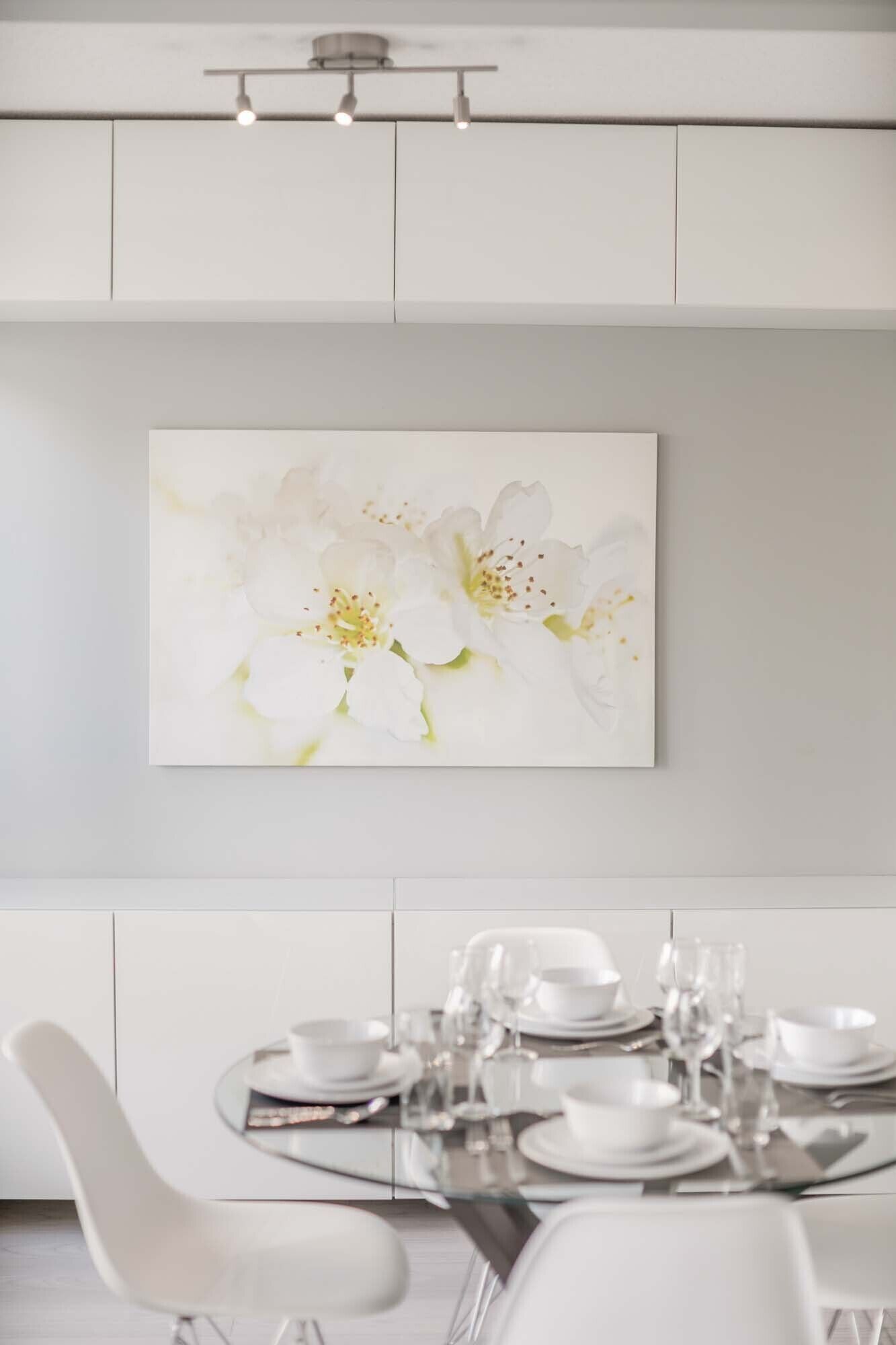 Zona pranzo con tavolo imbandito per sei persone, sedie bianche e un quadro appeso al muro raffigurante fiori bianchi.