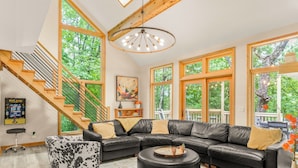 A spacious living room with a black leather sectional sofa, a round ottoman, and a staircase with wooden railings. - Newly Rebuilt Home - Hot Tub - Sauna - Sleeps 14 (Wintergreen Resort)