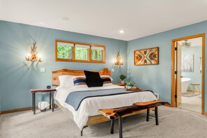 Un dormitorio con cama, mesitas de noche, un banco, una lámpara de pared y una ventana con persianas.