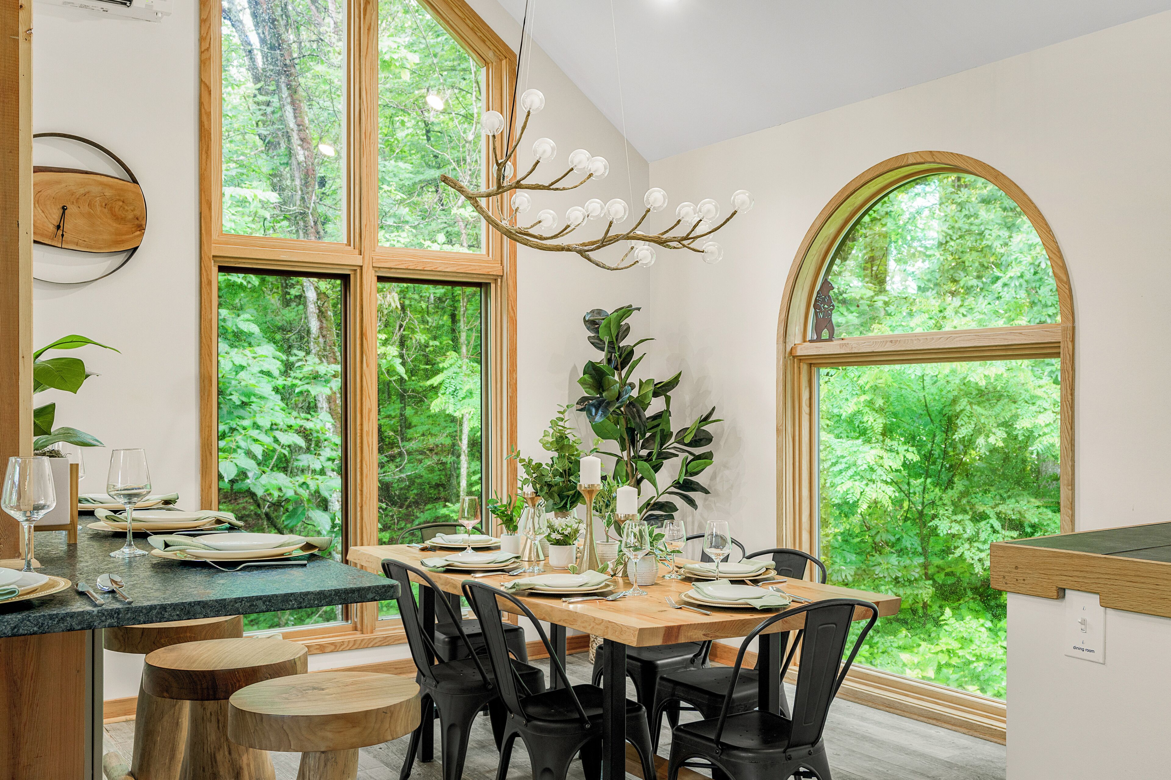 Comedor con mesa de madera dispuesta para seis personas, sillas negras, ventanas amplias con vista a la vegetación y una lámpara colgante original.