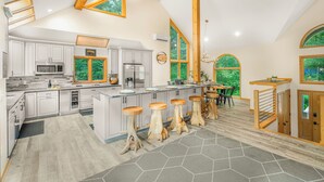 A spacious kitchen with a large island, wooden stools, and a view of greenery through large windows. - Newly Rebuilt Home - Hot Tub - Sauna - Sleeps 14 (Wintergreen Resort)